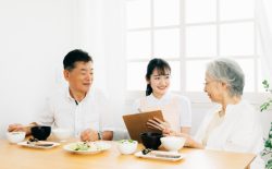 食事中の高齢夫婦と、介護士の女性がクリップボードを持って話しかけている様子。