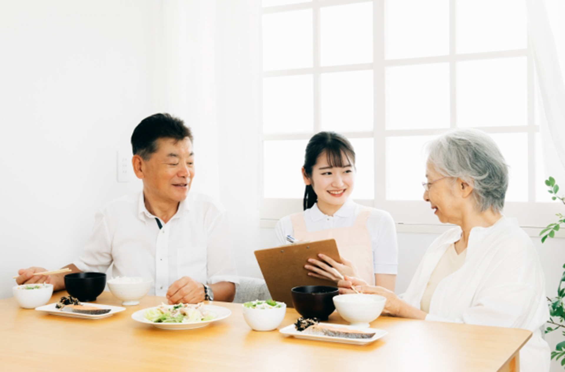 食事中の高齢夫婦と、介護士の女性がクリップボードを持って話しかけている様子。