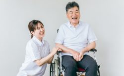 車椅子に座った笑顔の高齢男性と、その横にしゃがみ込み、笑いかける介護士の若い女性。