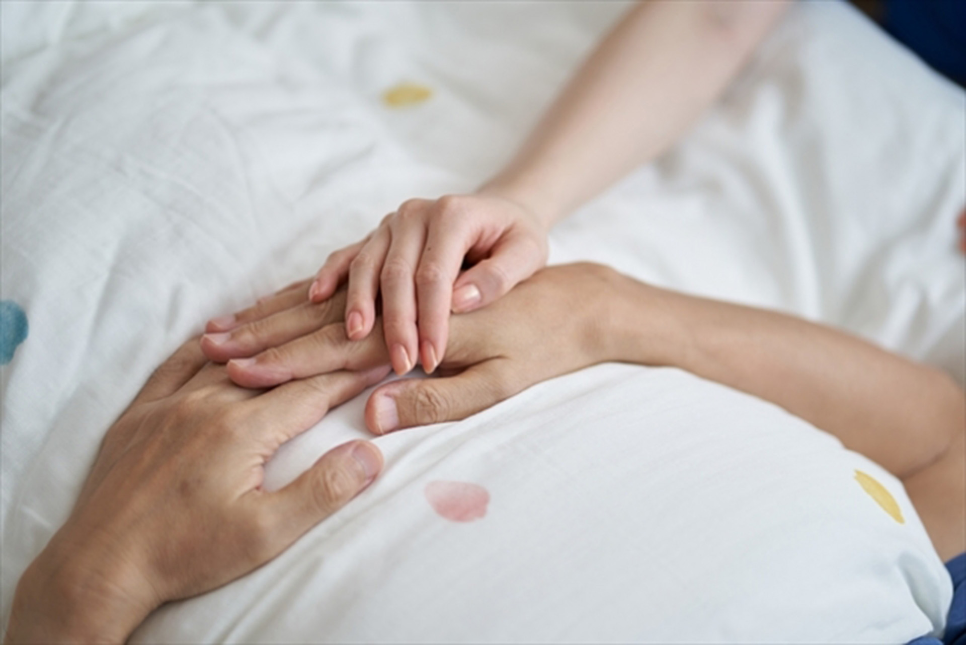 ベッドに寝ている老人のの手と手を重ねる女性の様子
